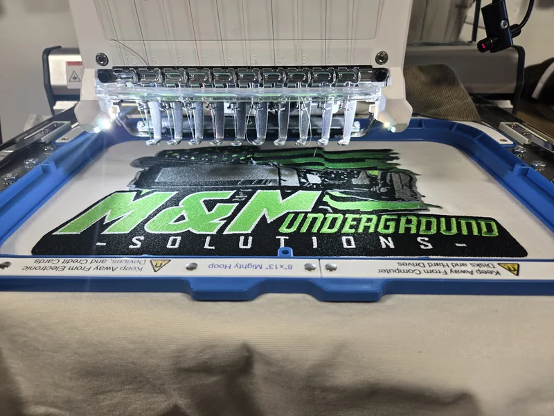 Multi-needle embroidery machine stitching the M&M Underground Solutions logo in green and white