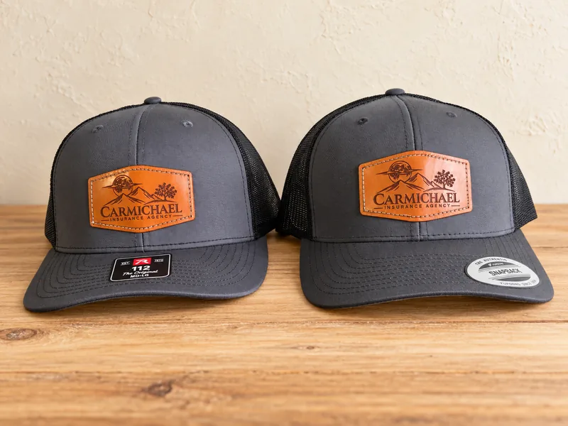 Two charcoal and black trucker hats with laser-engraved leather Carmichael Insurance Agency patches